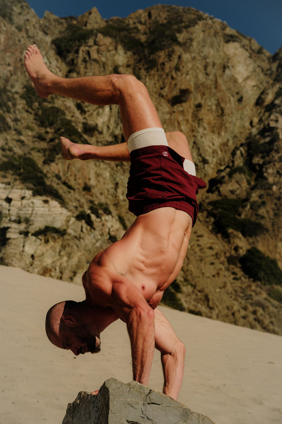 A handstand on a rock above the beach, wearing Wayve shorts