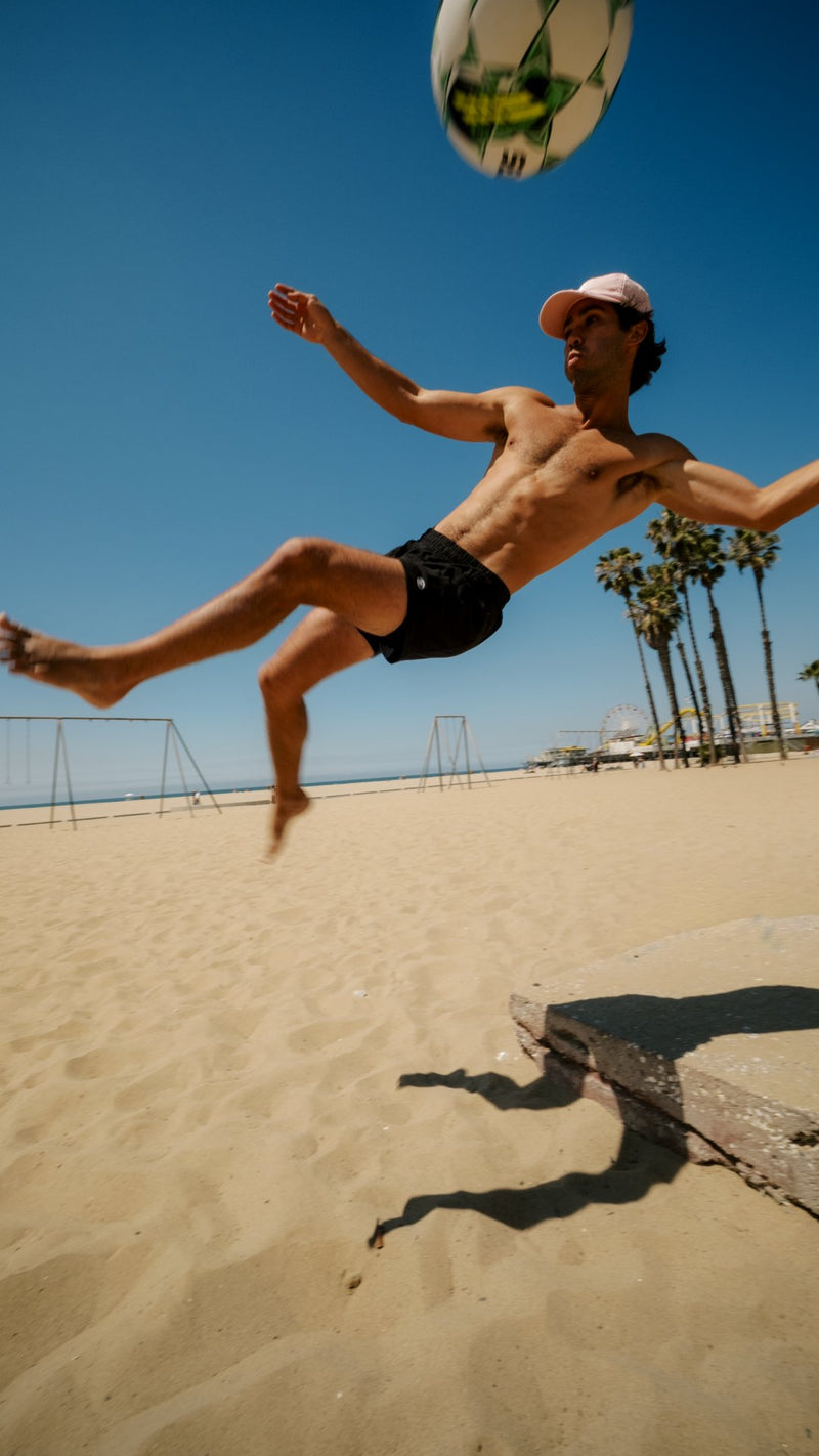 Yahli mid-air playing football on the sand in Wayve shorts