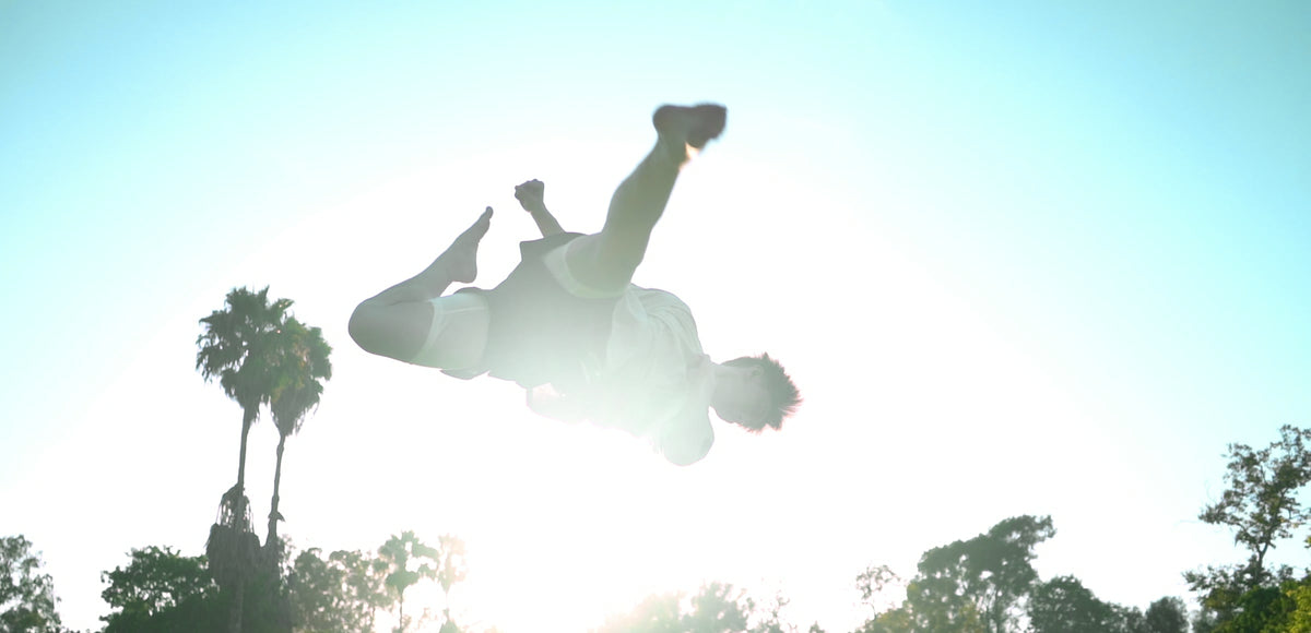 A man mid-backflip against the sun, palm trees below